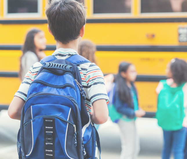 A boy with a blue backpack stands in front of a yellow school bus