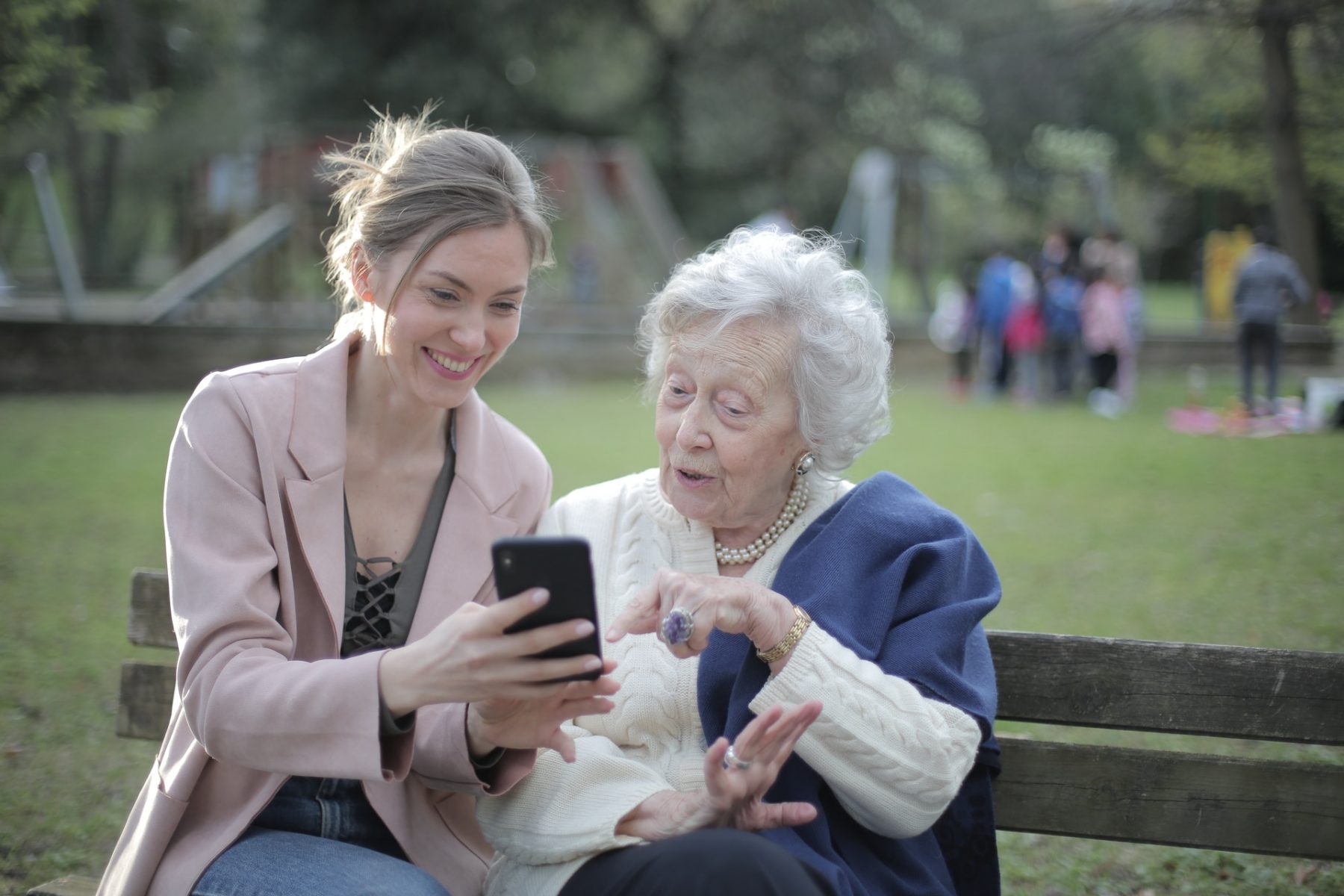 middle aged women showing an elderly women something on her phone while sitting on a park bench